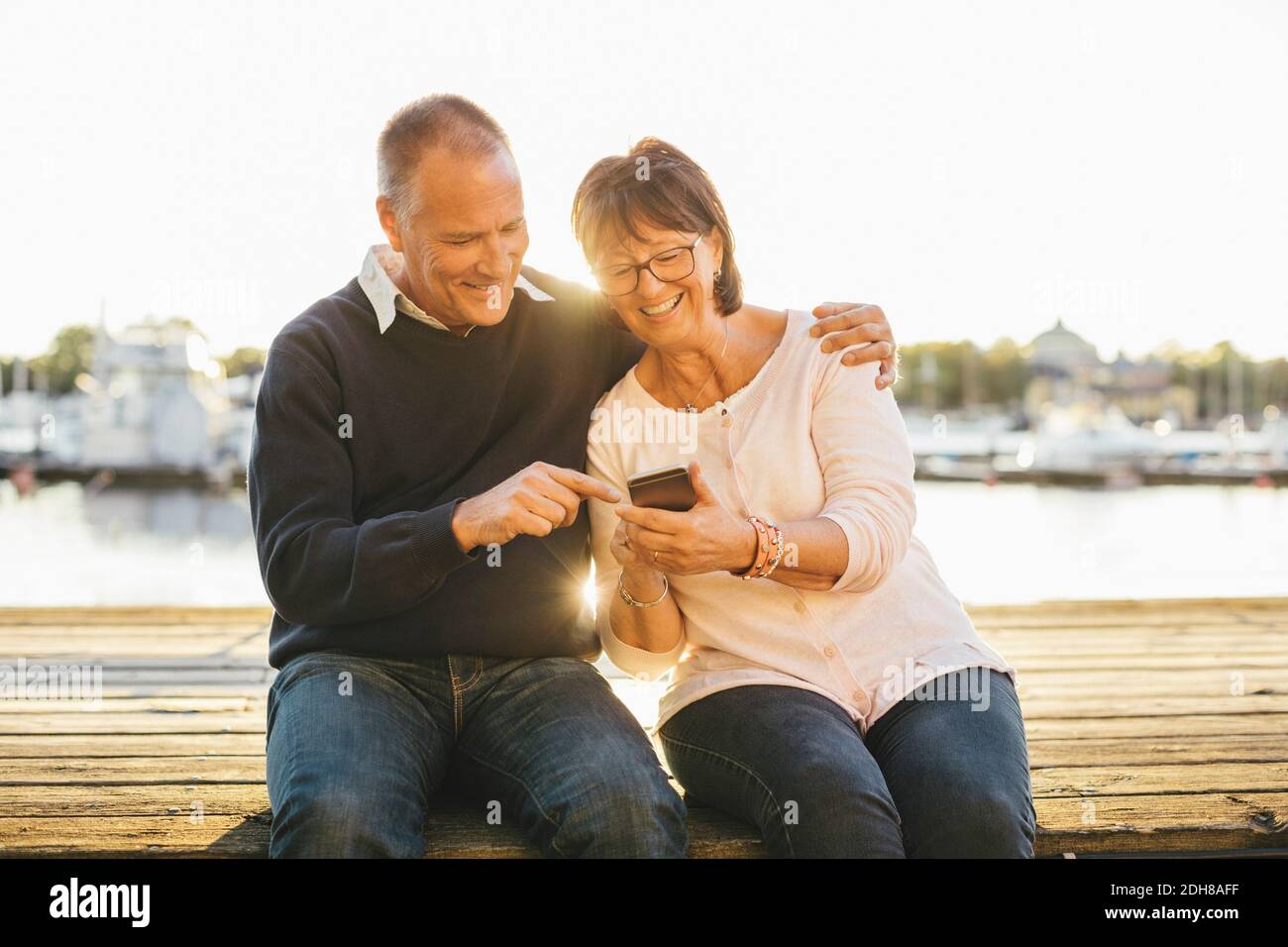 Happy senior couple using mobile phone while sitting on pier Stock ...