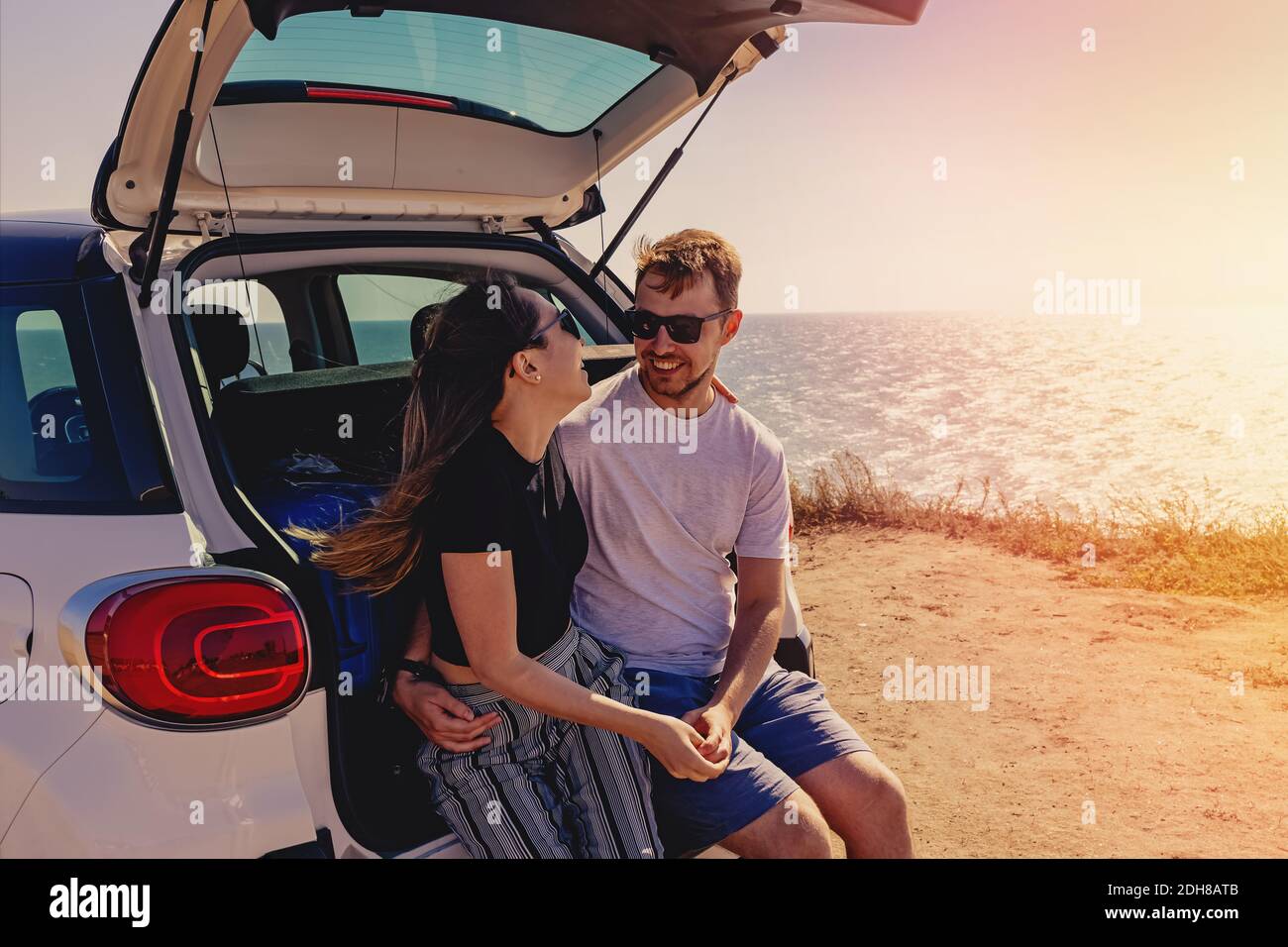 Happy young couple sitting on the trunk of the car near the sea. Summer travel, road trip Stock Photo