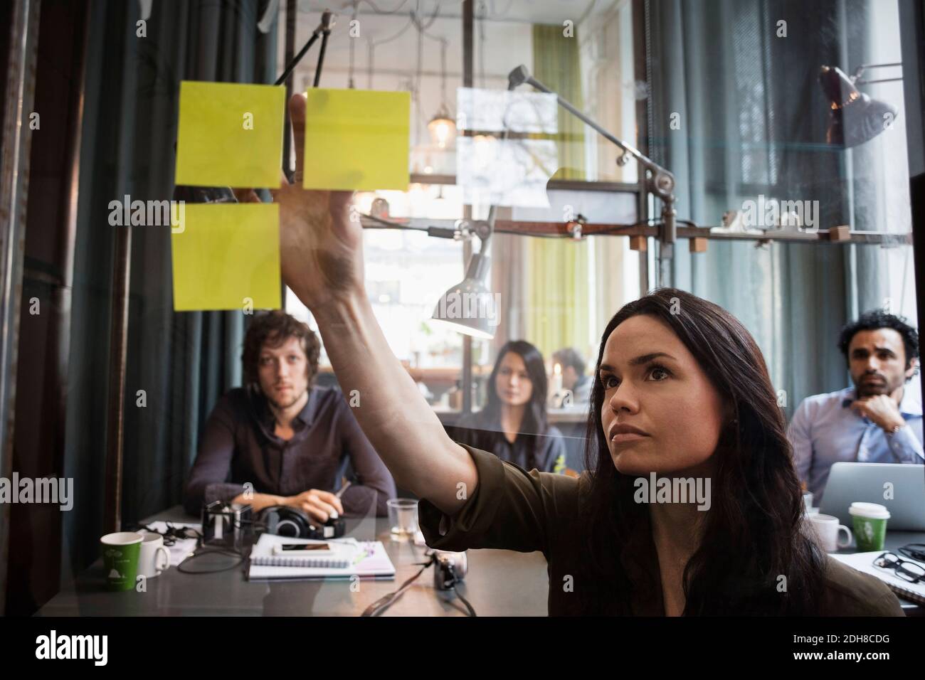 Creative business people looking at female colleague sticking adhesive note on window seen through glass Stock Photo