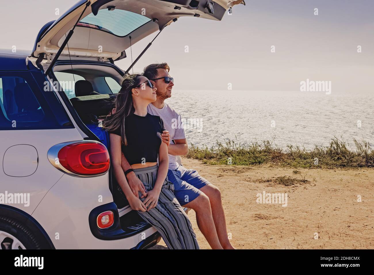 Happy young couple sitting on the trunk of the car near the sea. Summer travel, road trip Stock Photo