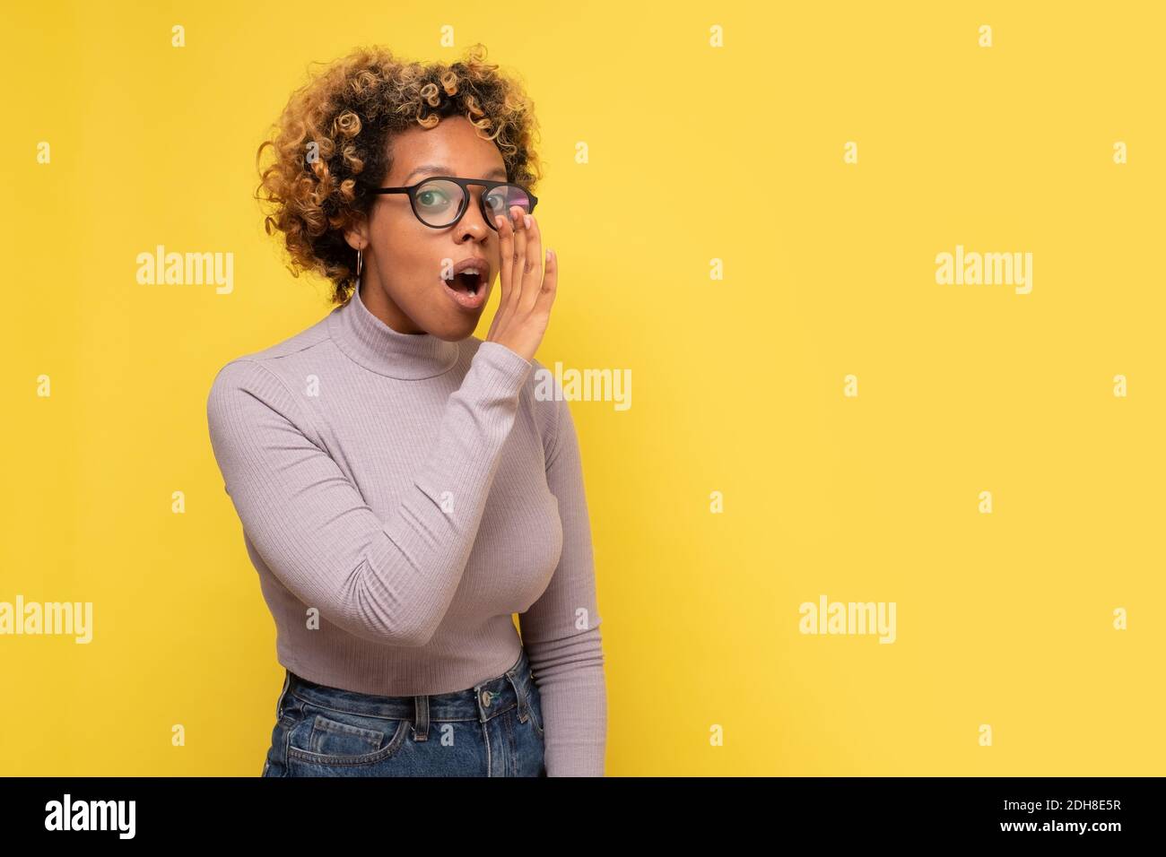 Young african american woman with hand on mouth telling secret rumor. Studio shot on yellow wall. Stock Photo