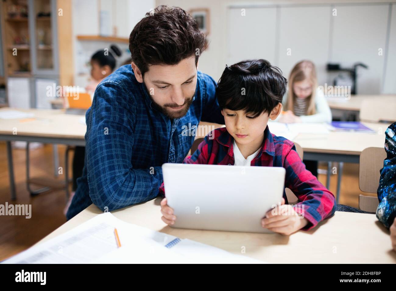 Teacher assisting male student in using digital tablet while sitting in classroom Stock Photo
