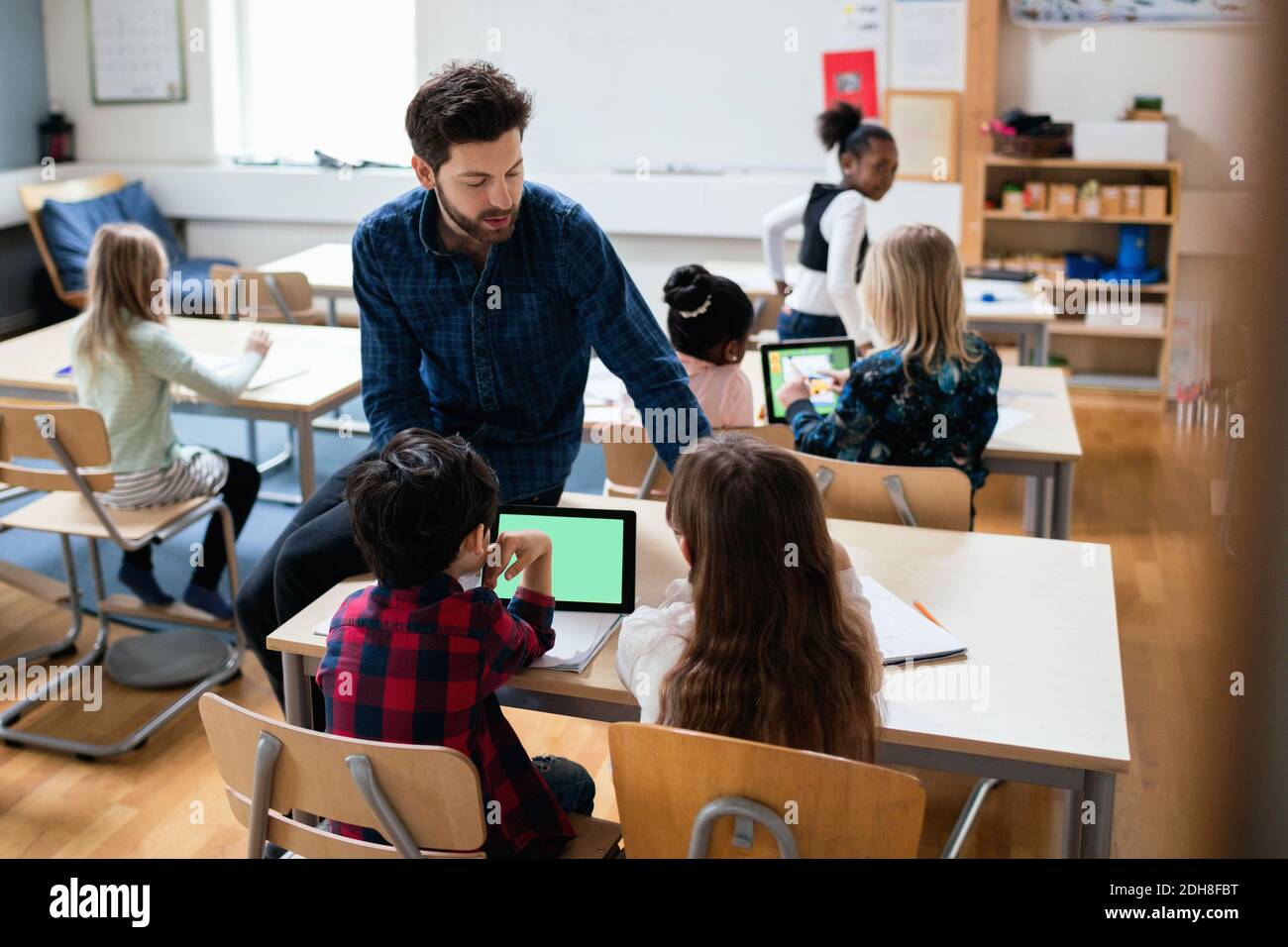 Teacher sitting on desk while explaining students using digital tablet in classroom Stock Photo