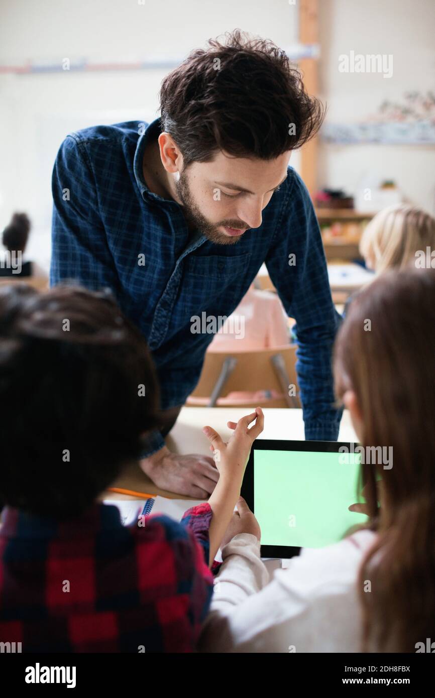 Male teacher looking at student using digital tablet while sitting by boy in classroom Stock Photo