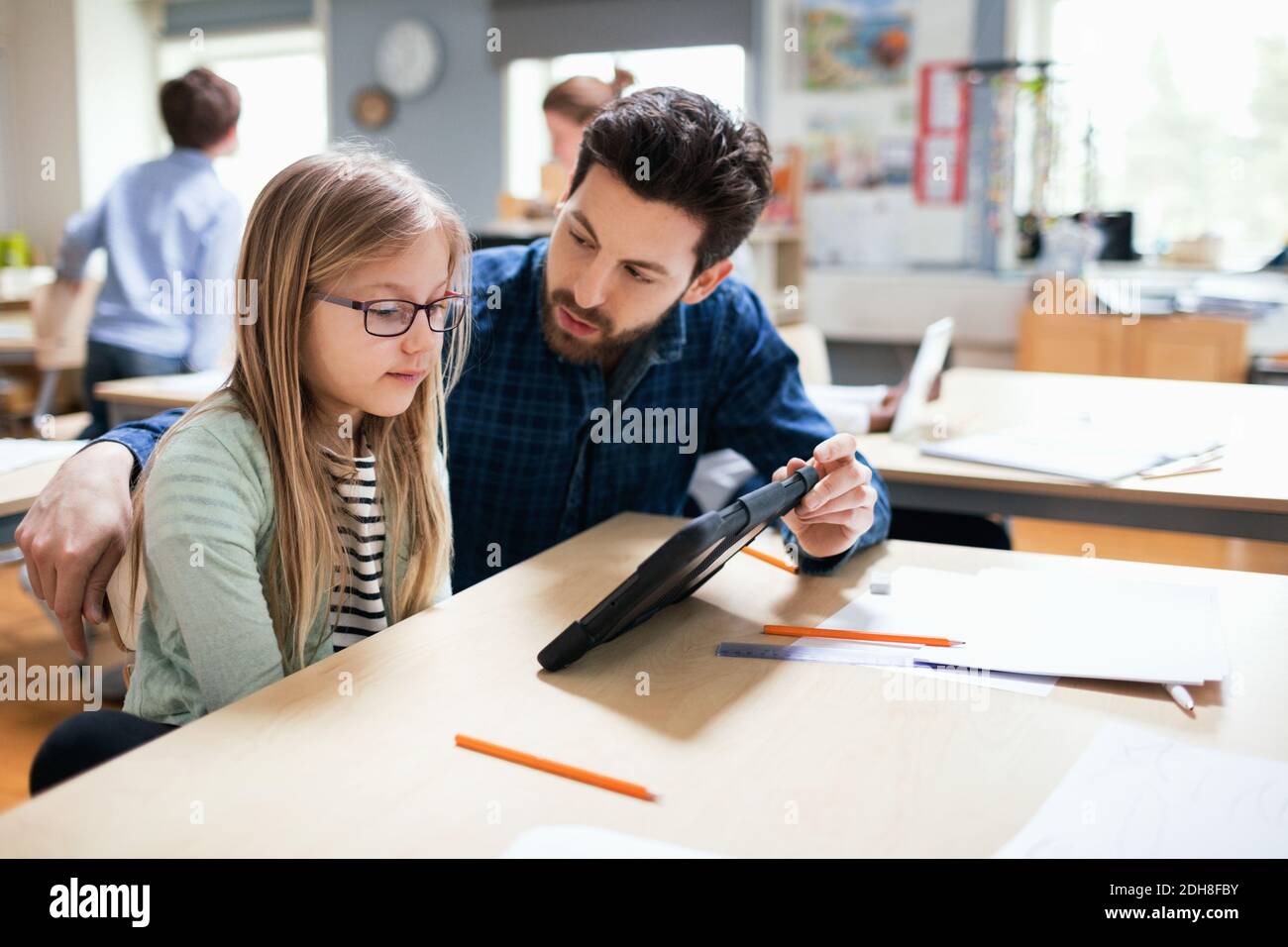 Teacher discussing with student while showing digital tablet at desk in classroom Stock Photo
