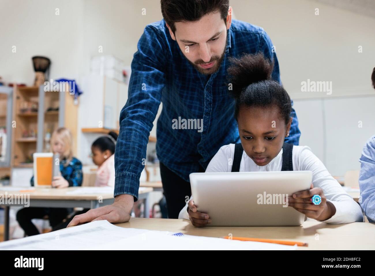 Teacher looking at girl using digital tablet in classroom Stock Photo