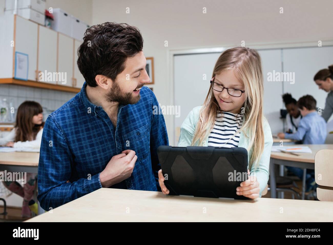 Smiling teacher looking at girl holding digital tablet at desk Stock Photo