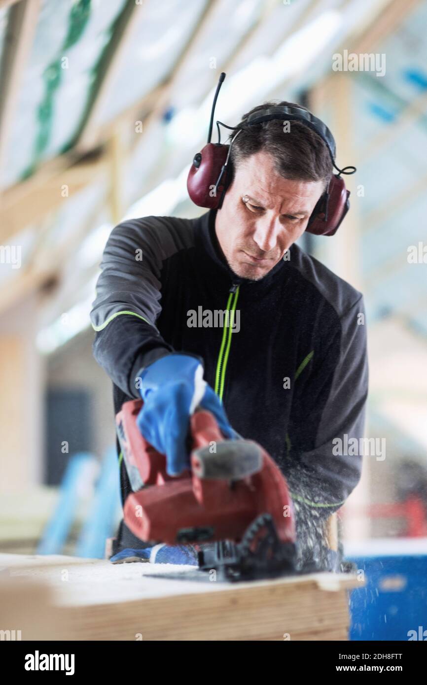Concentrated manual worker wearing ear protectors sawing wooden planks ...