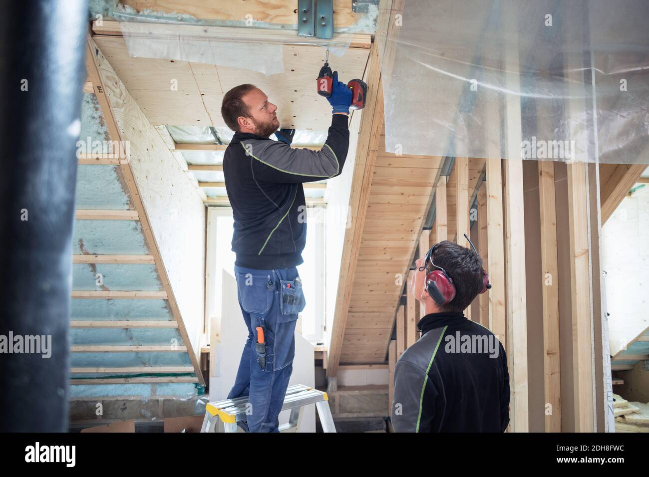 Coworker looking at carpenter drilling on roof beam in construction site Stock Photo