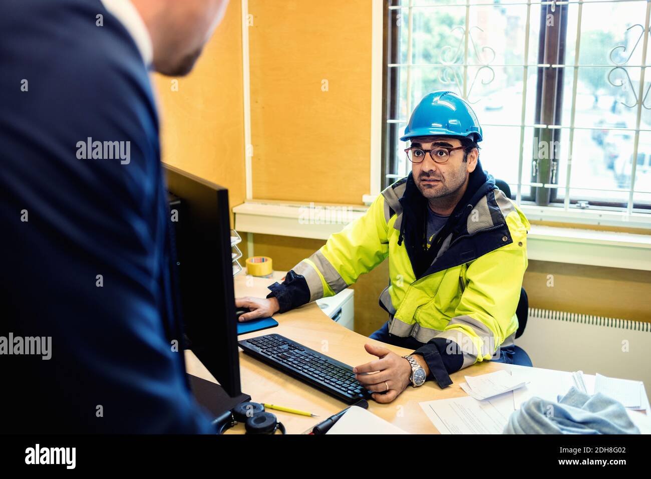 Serious construction worker in protective wear talking to man at desk Stock Photo