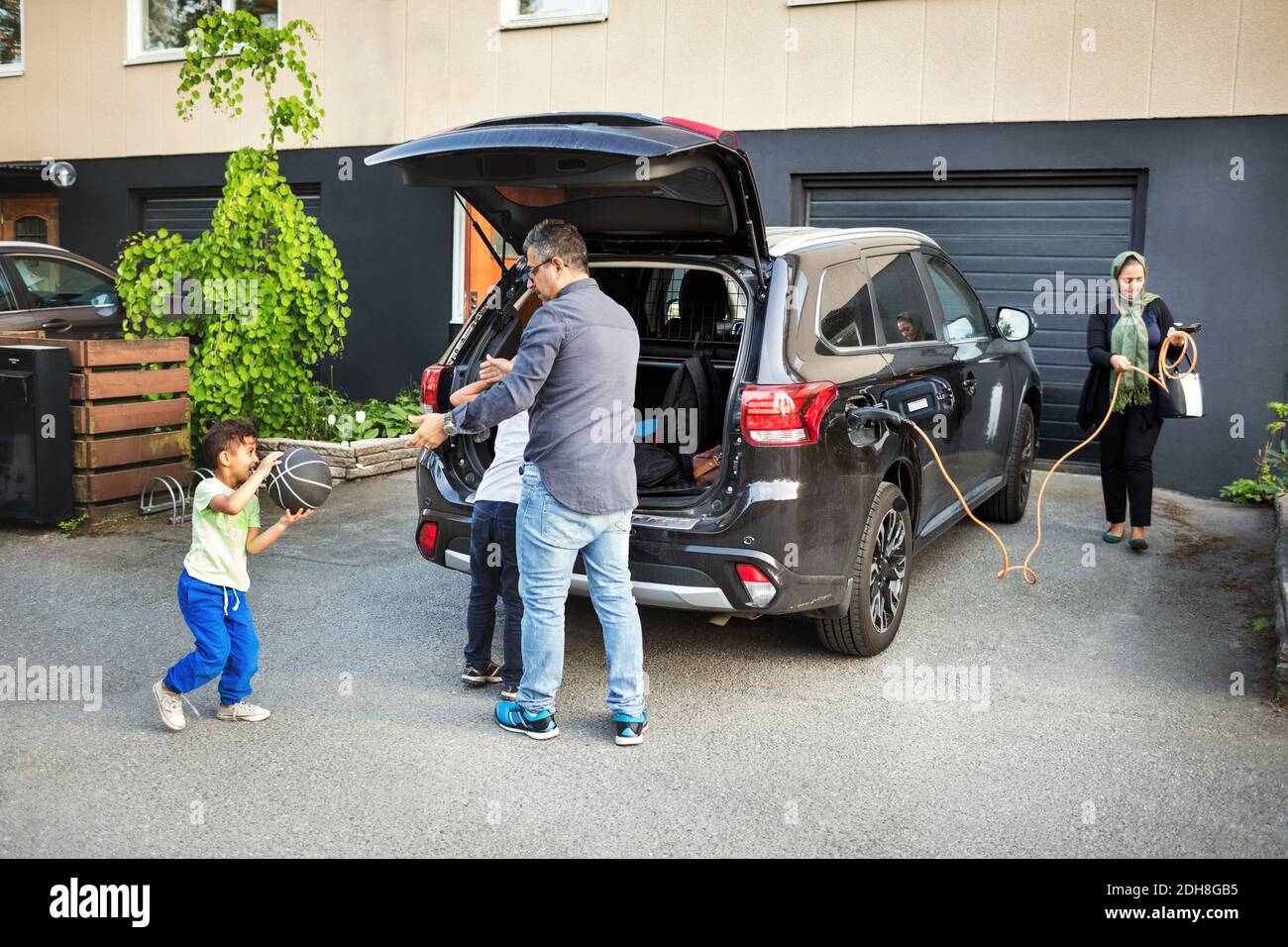 Father playing with children while mother rolling electric charger by car Stock Photo