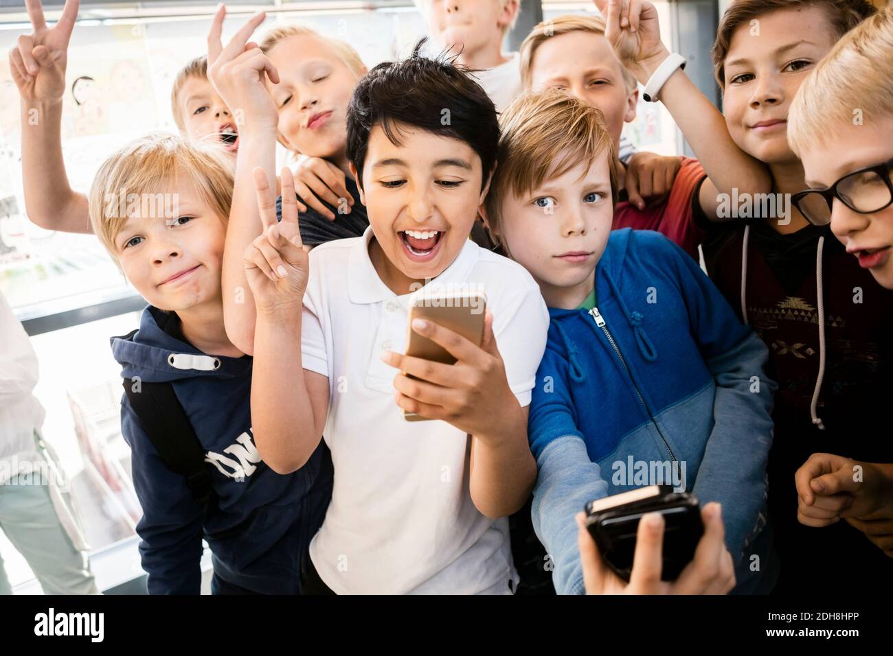 Happy school kids using mobile phone with peace sign in corridor Stock Photo