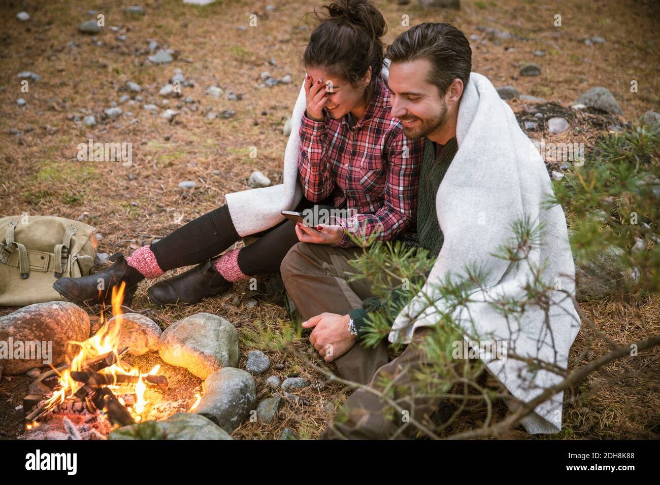 High angle view of happy couple wrapped in blanket while using mobile phone by fire pit at campsite Stock Photo