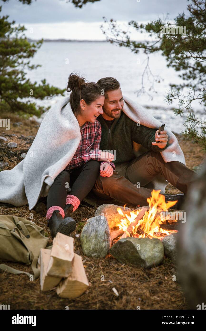 Couple wrapped in blanket using mobile phone while sitting by fire pit at campsite Stock Photo