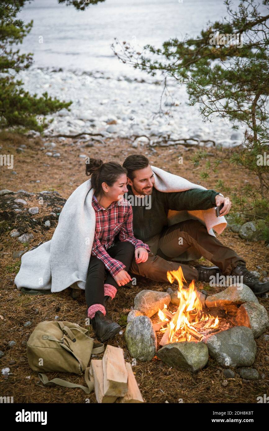 Couple wrapped in blanket taking selfie while sitting by fire pit at campsite Stock Photo