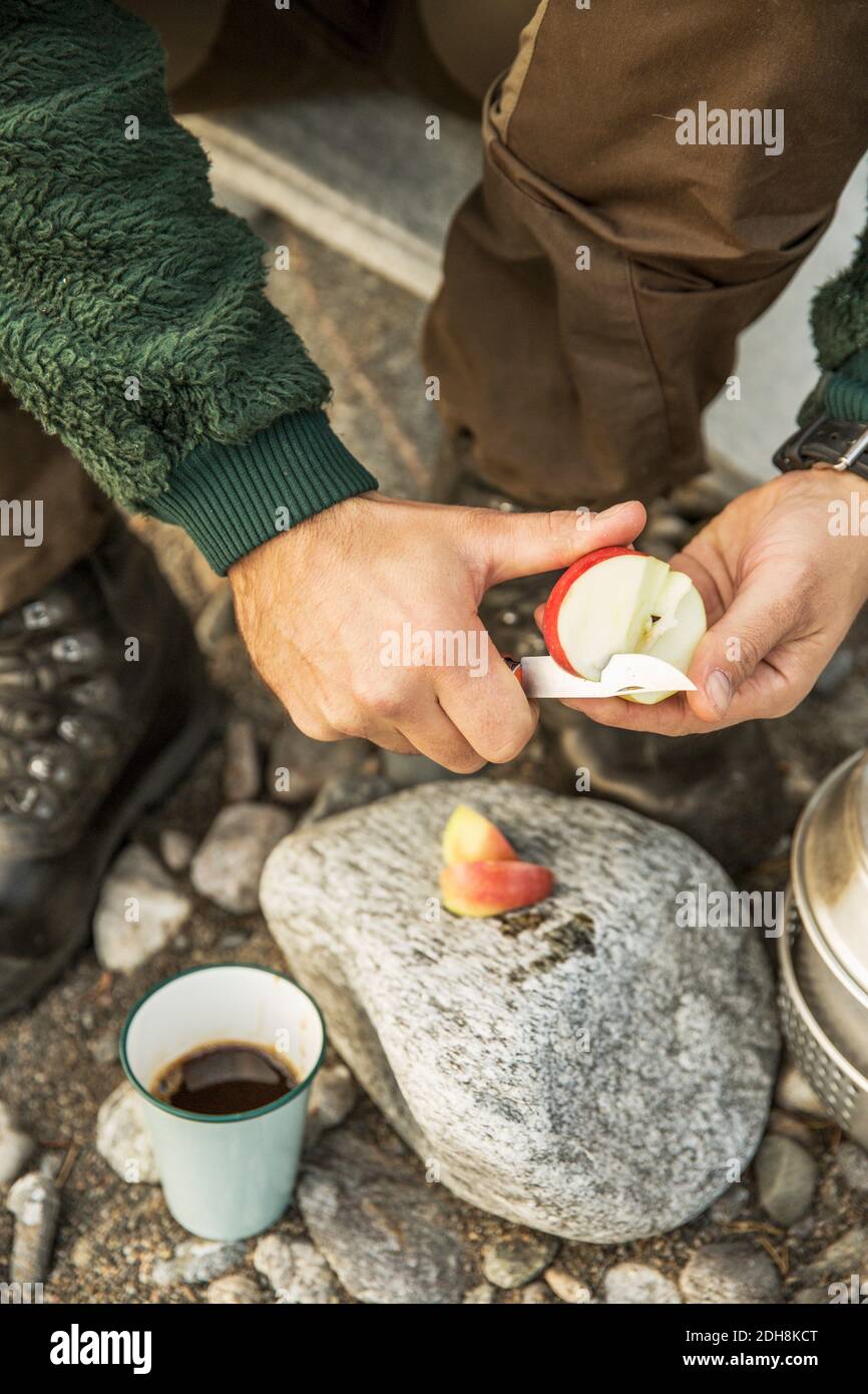 Midsection of man cutting apple over rock by coffee cup at camp Stock Photo