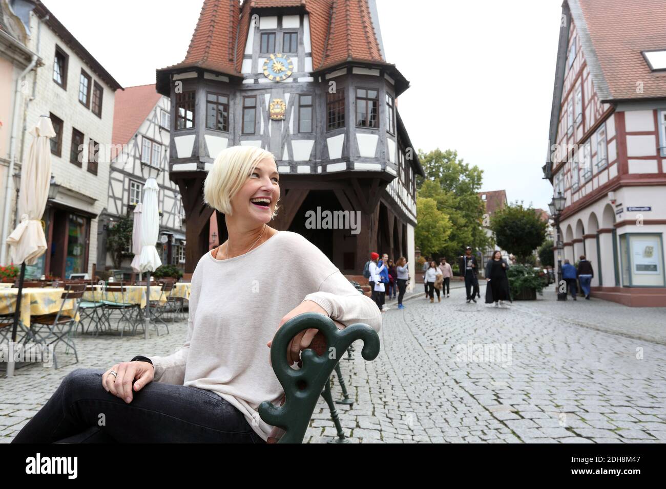 Young woman sitting on bench and smiling with town hall of Michelstadt in the background , Hessen, Germany. Stock Photo