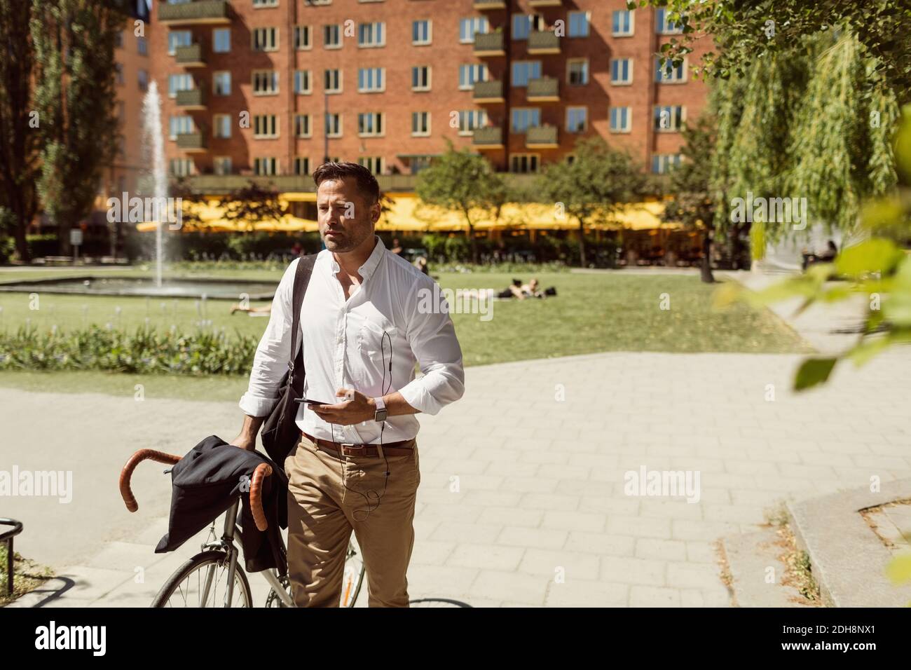 Thoughtful businessman holding bicycle and smart phone on footpath during summer Stock Photo