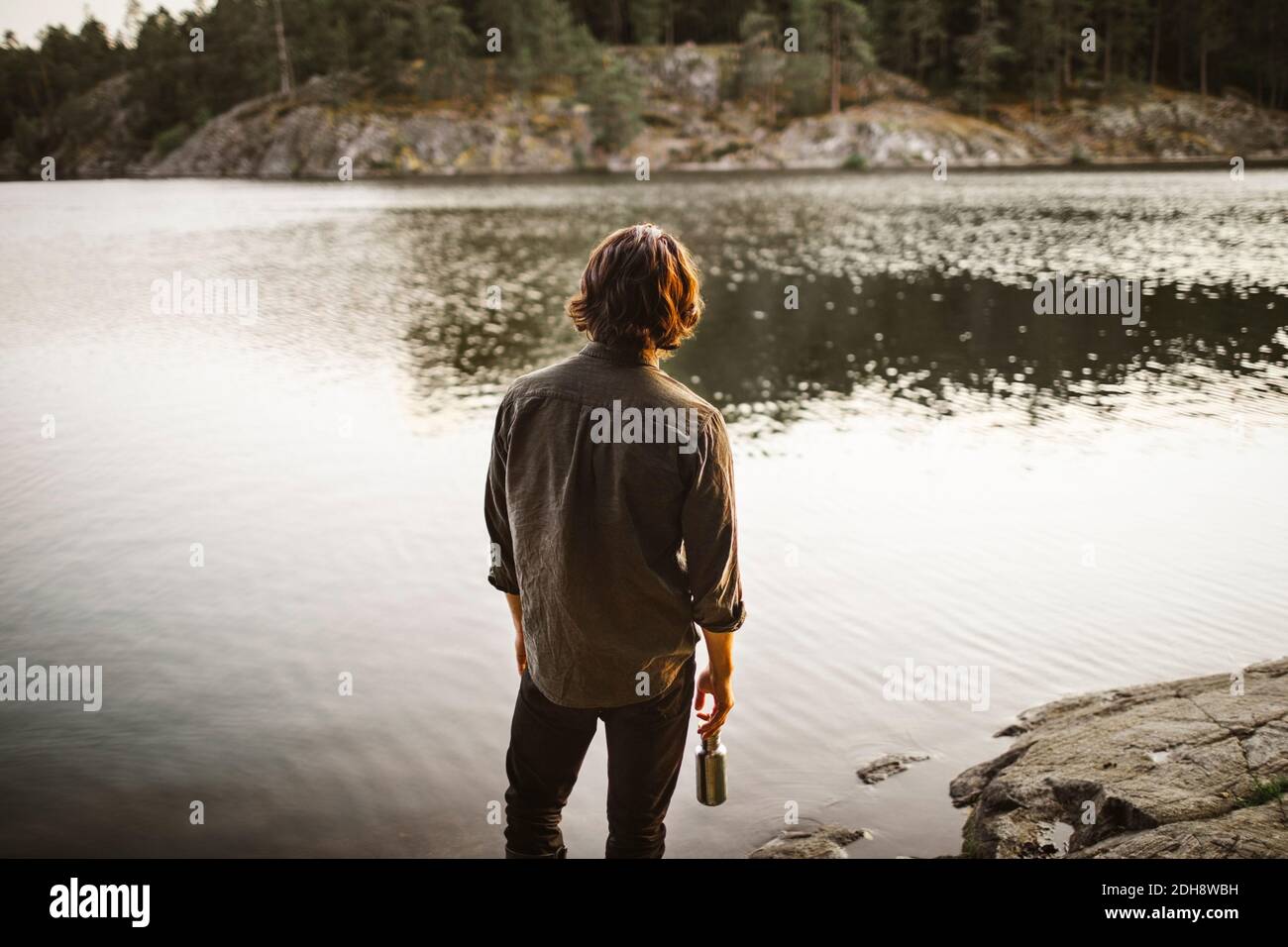 Rear view of young man standing by lake in forest during vacation Stock Photo