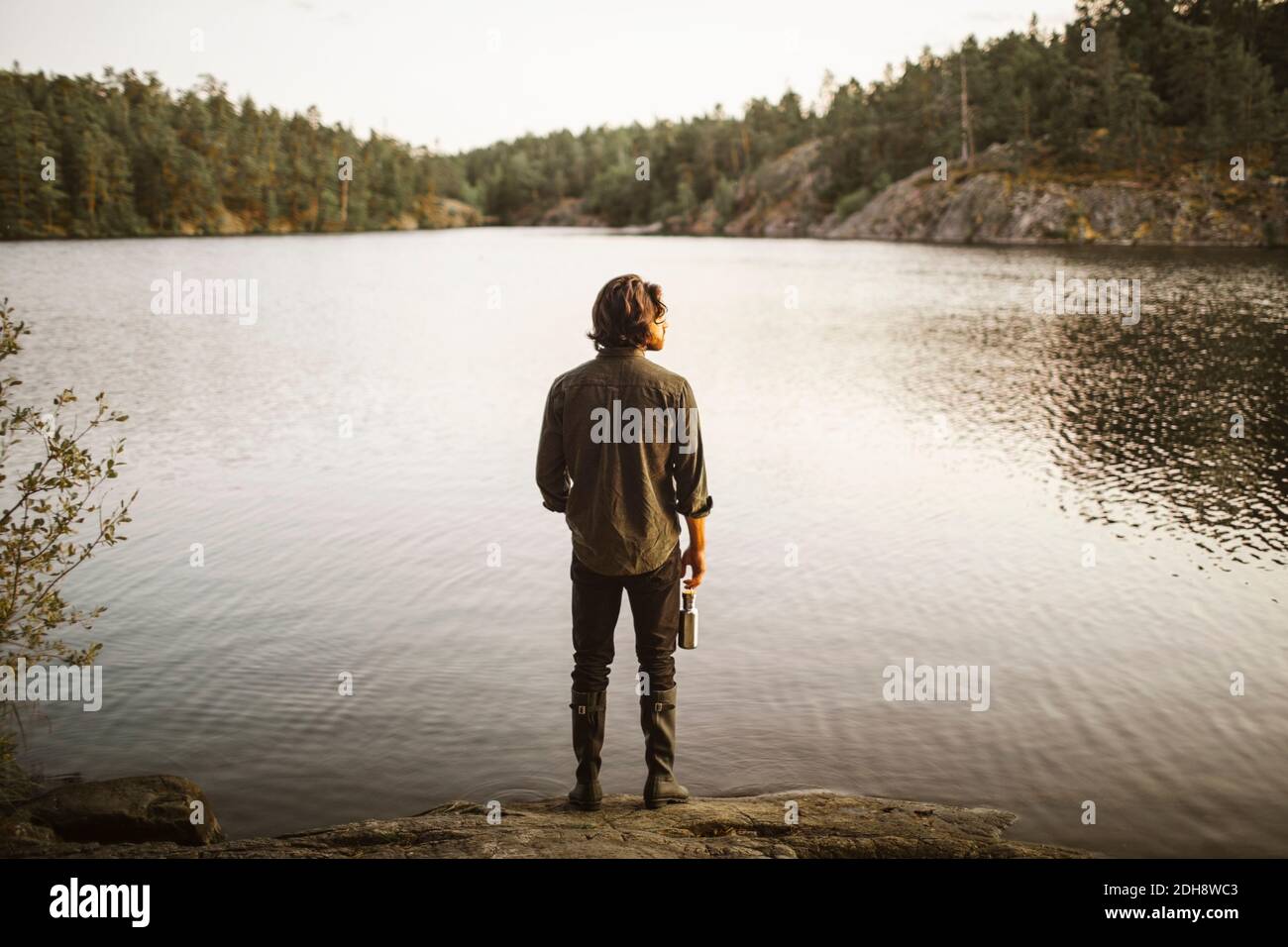 Rear view of man with bottle standing by lake in forest during vacation Stock Photo