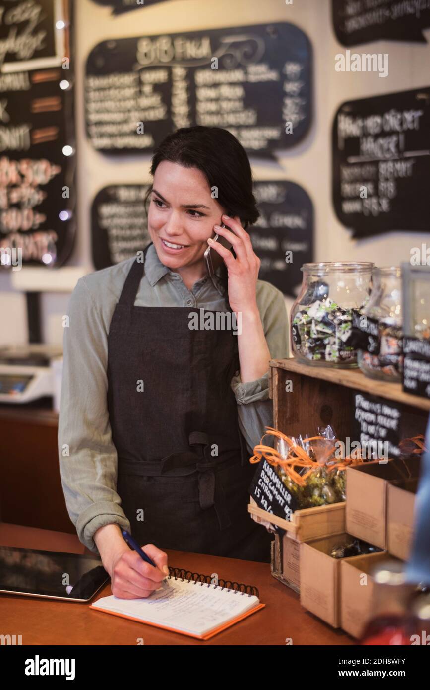 Smiling female owner using phone while writing in note pad at store ...