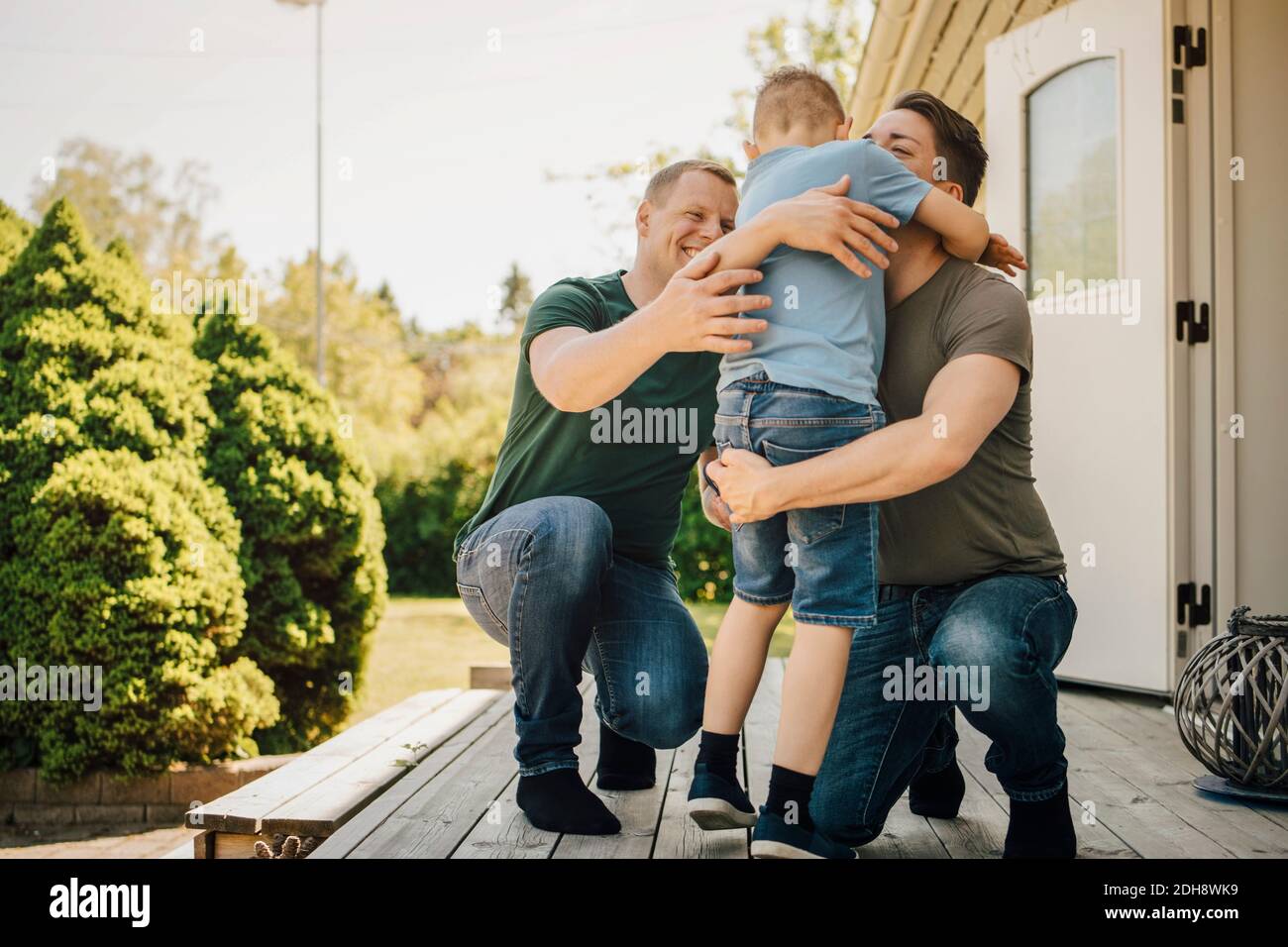 Homosexual fathers embracing son while crouching on patio during sunny day Stock Photo