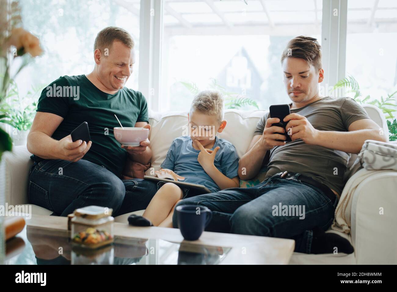 Smiling father with smart phone looking at son using digital tablet on sofa at home Stock Photo