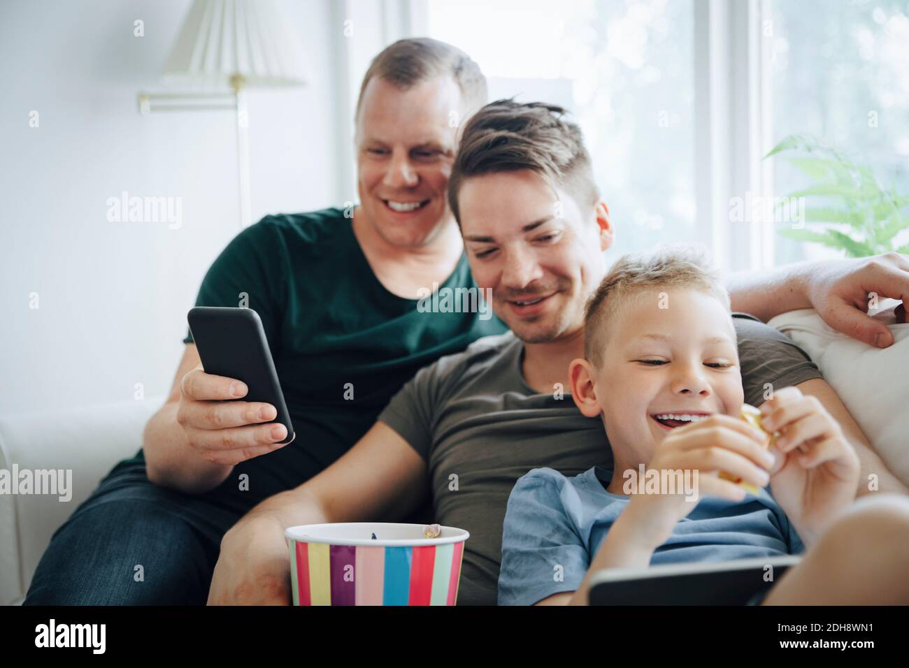 Smiling fathers and son using technology while eating popcorn on sofa at home Stock Photo