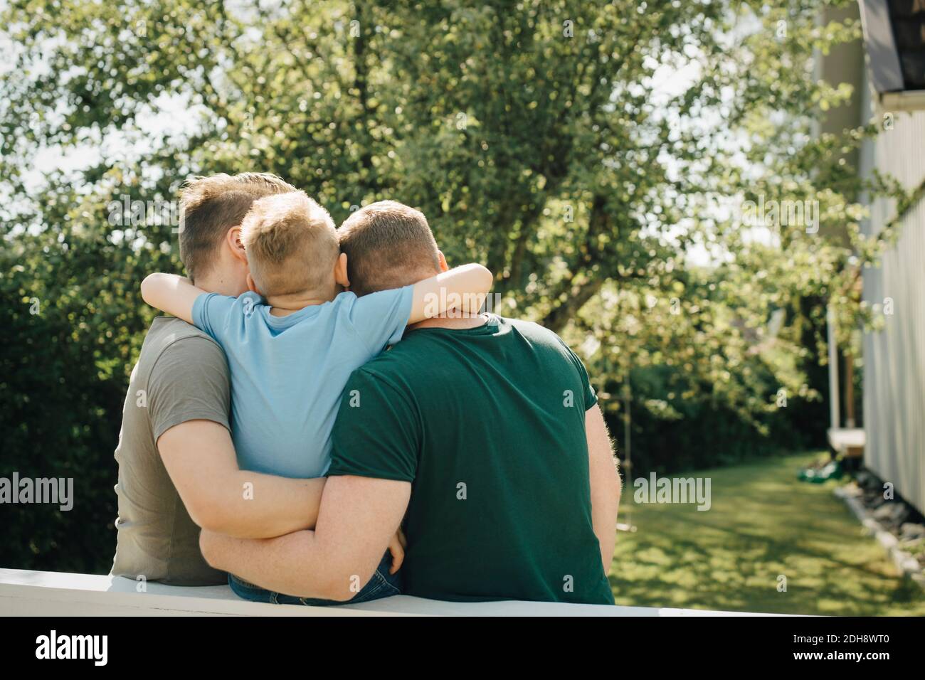 Rear view of son with arm around fathers sitting on fence in yard Stock Photo