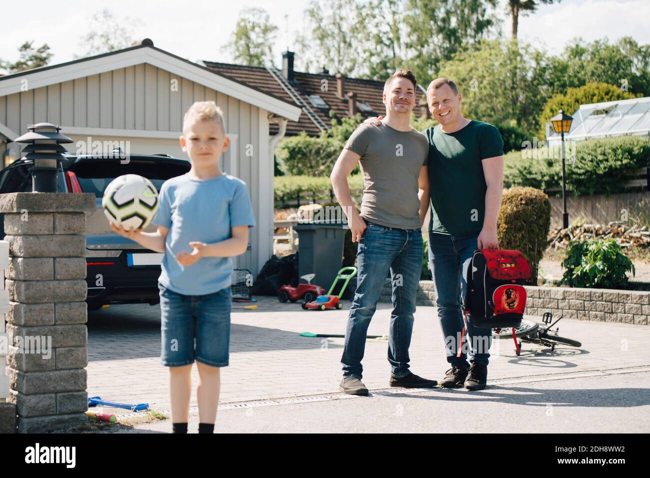 Portrait of homosexual fathers looking at son holding soccer ball outdoors during sunny day Stock Photo