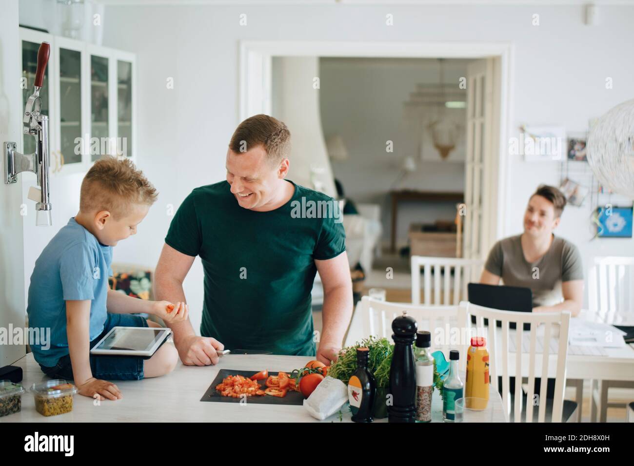 Smiling father talking to son holding tomato while using digital tablet on kitchen counter Stock Photo