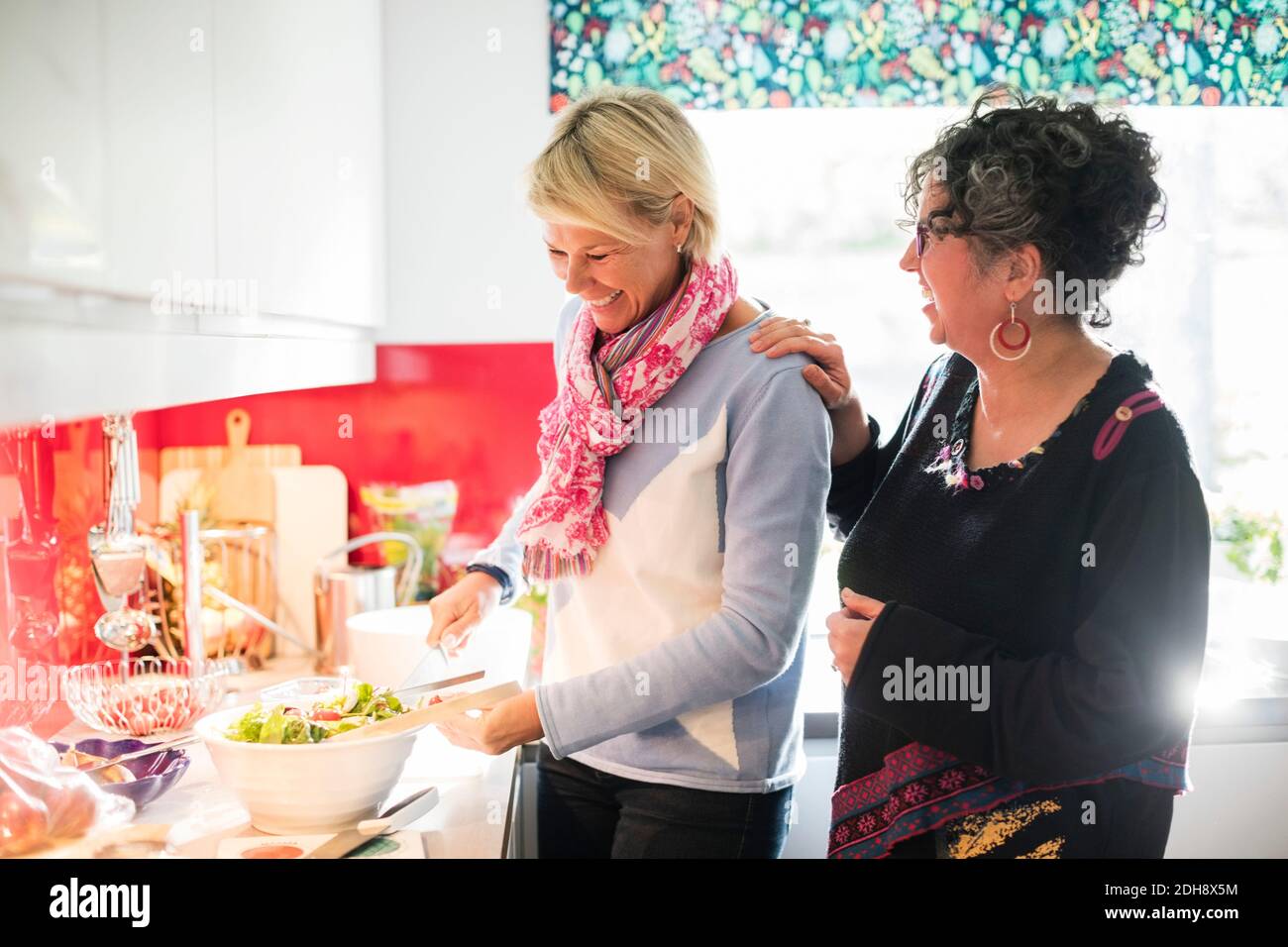 Happy friend talking to woman while preparing salad in brightly lit kitchen Stock Photo