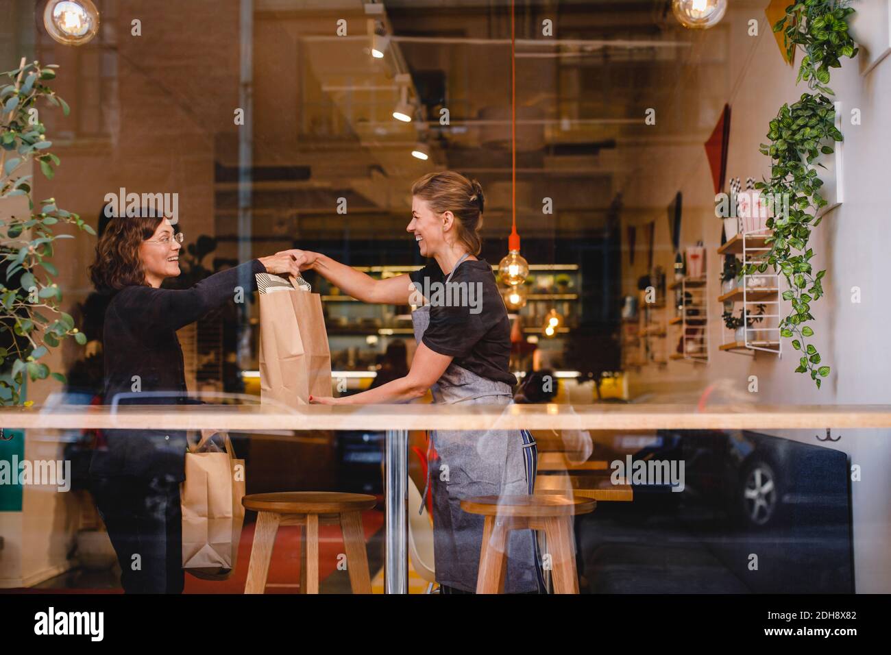 Side view of smiling saleswoman giving shopping bag to female customer at checkout counter seen through glass window Stock Photo