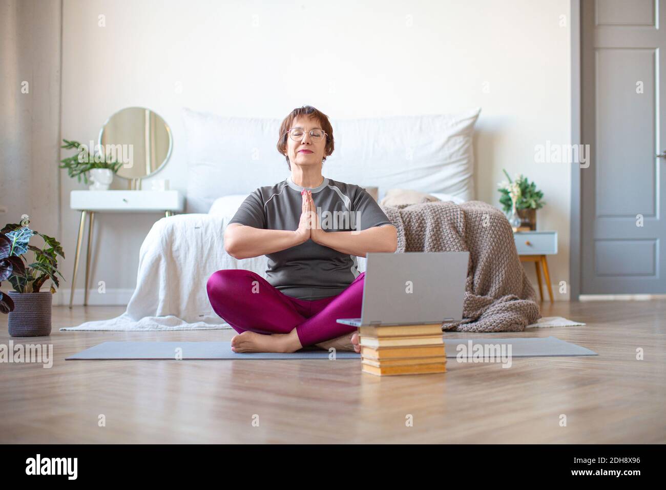 An elderly woman meditates in lotus pose at home in front of a laptop monitor. The concept of a healthy and active lifestyle in old age. Stock Photo