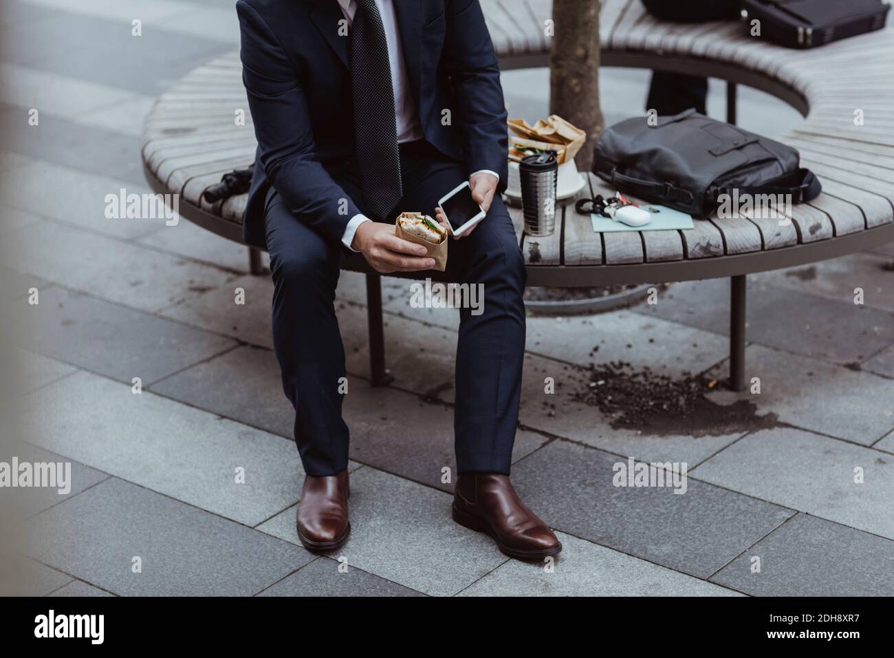 Low section of entrepreneur with smart phone and sandwich sitting in city Stock Photo