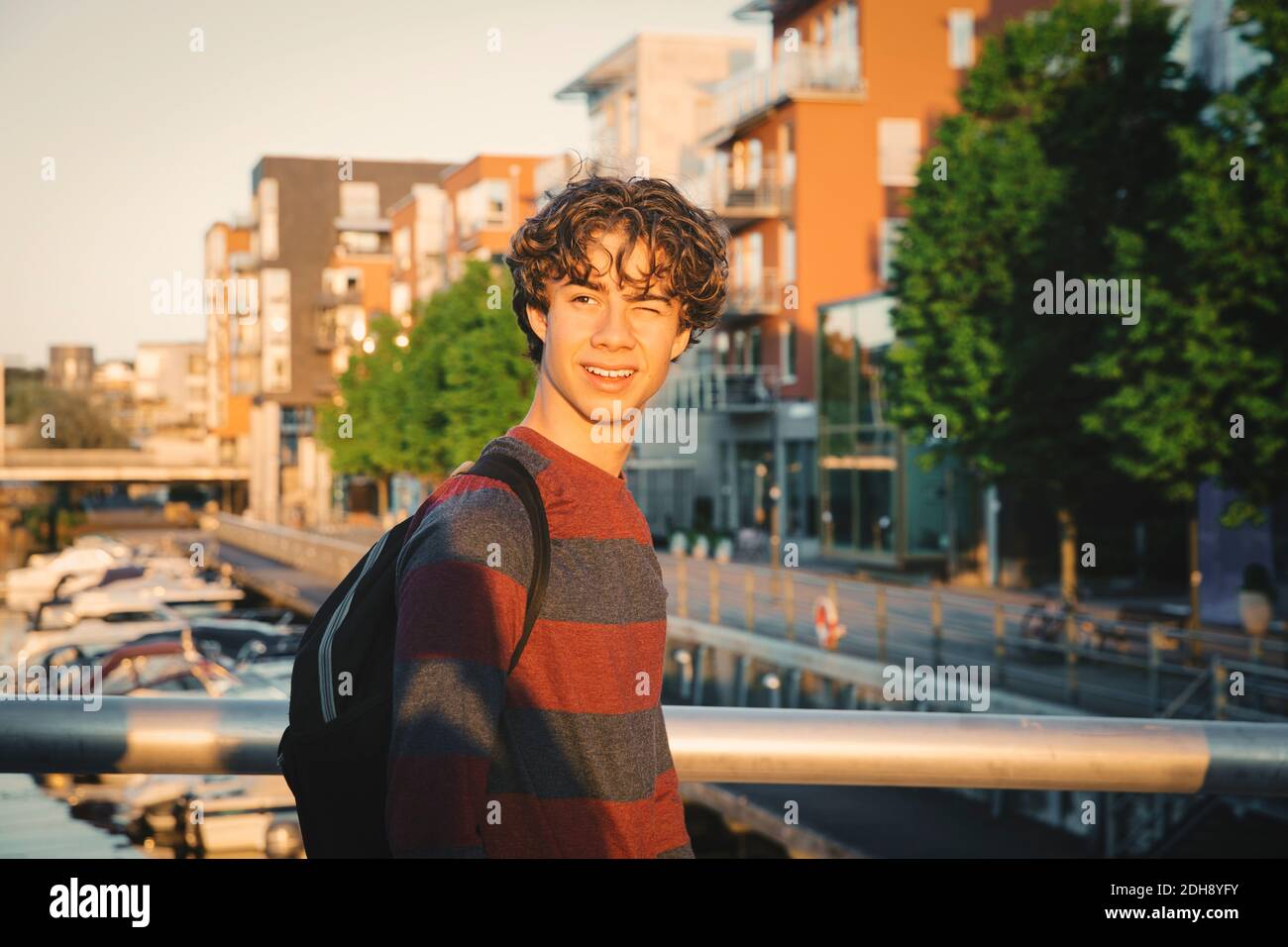 Smiling teenage boy winking on bridge in city during sunset Stock Photo