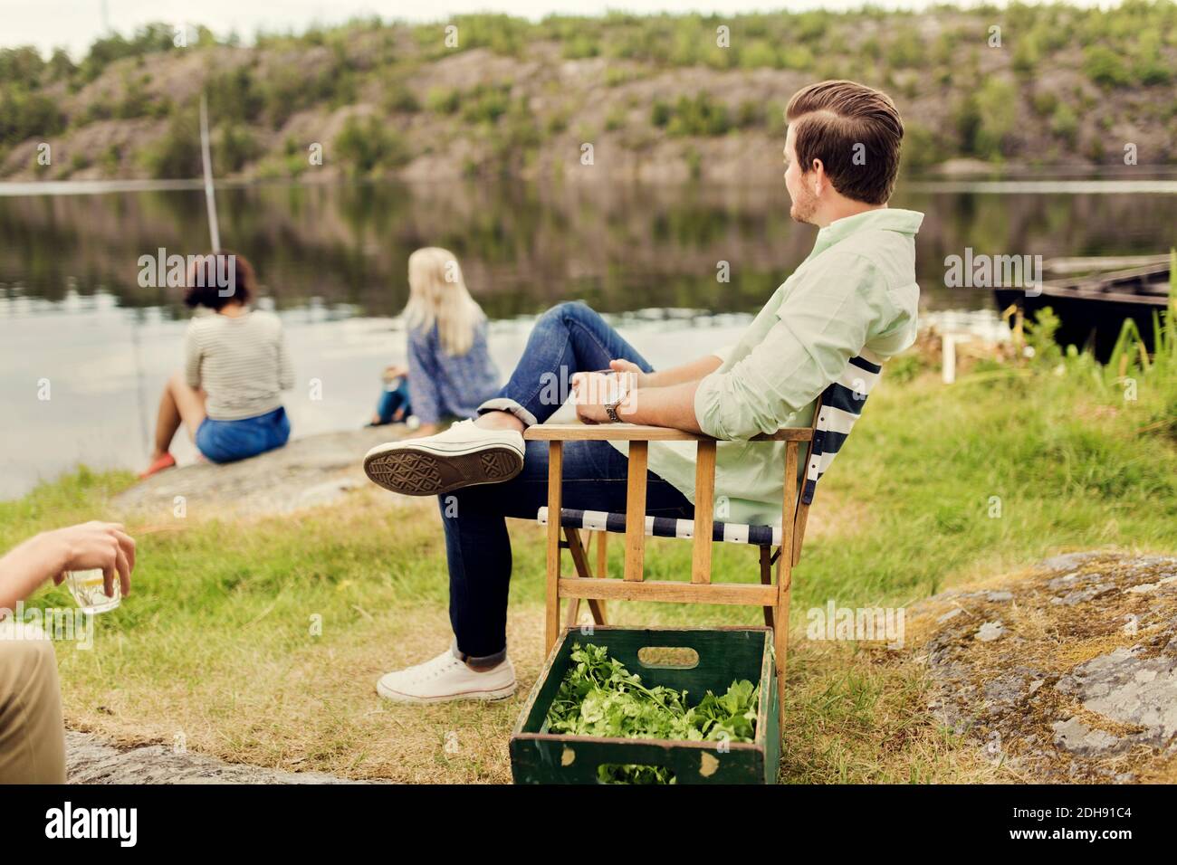 Men having drink while looking at female friends fishing on lakeshore Stock Photo