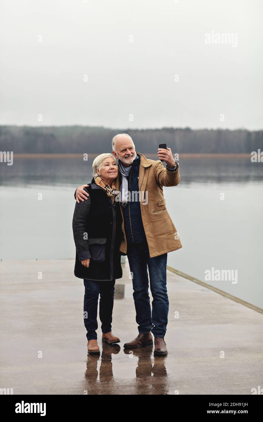 Full length of senior couple taking selfie at lakeside against clear sky Stock Photo
