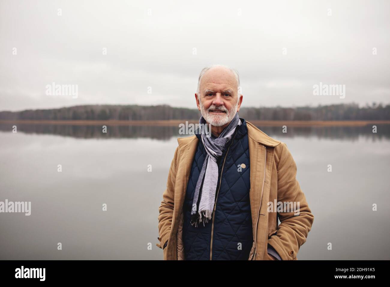 Portrait of smiling senior man with hands in pockets standing against lake Stock Photo