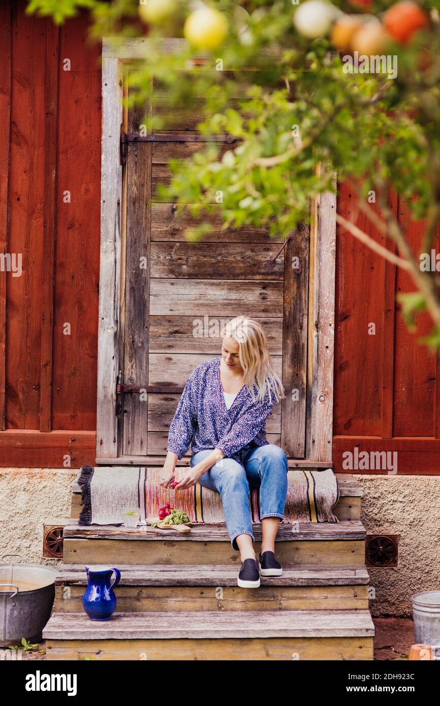 Full length of woman cutting radishes while sitting on steps outside wooden cottage Stock Photo