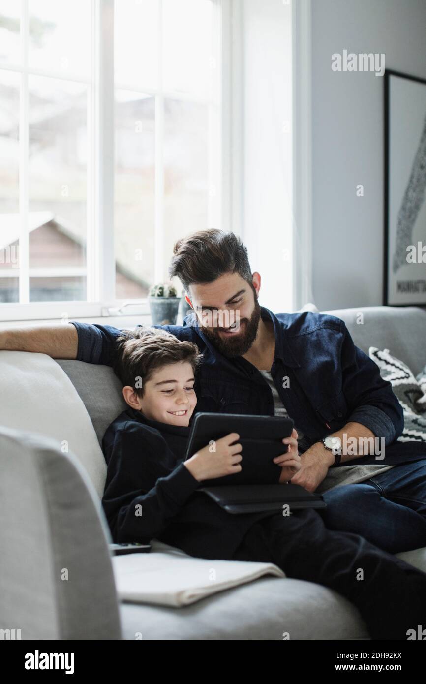 Happy father looking at boy using digital tablet while sitting on sofa in living room Stock Photo
