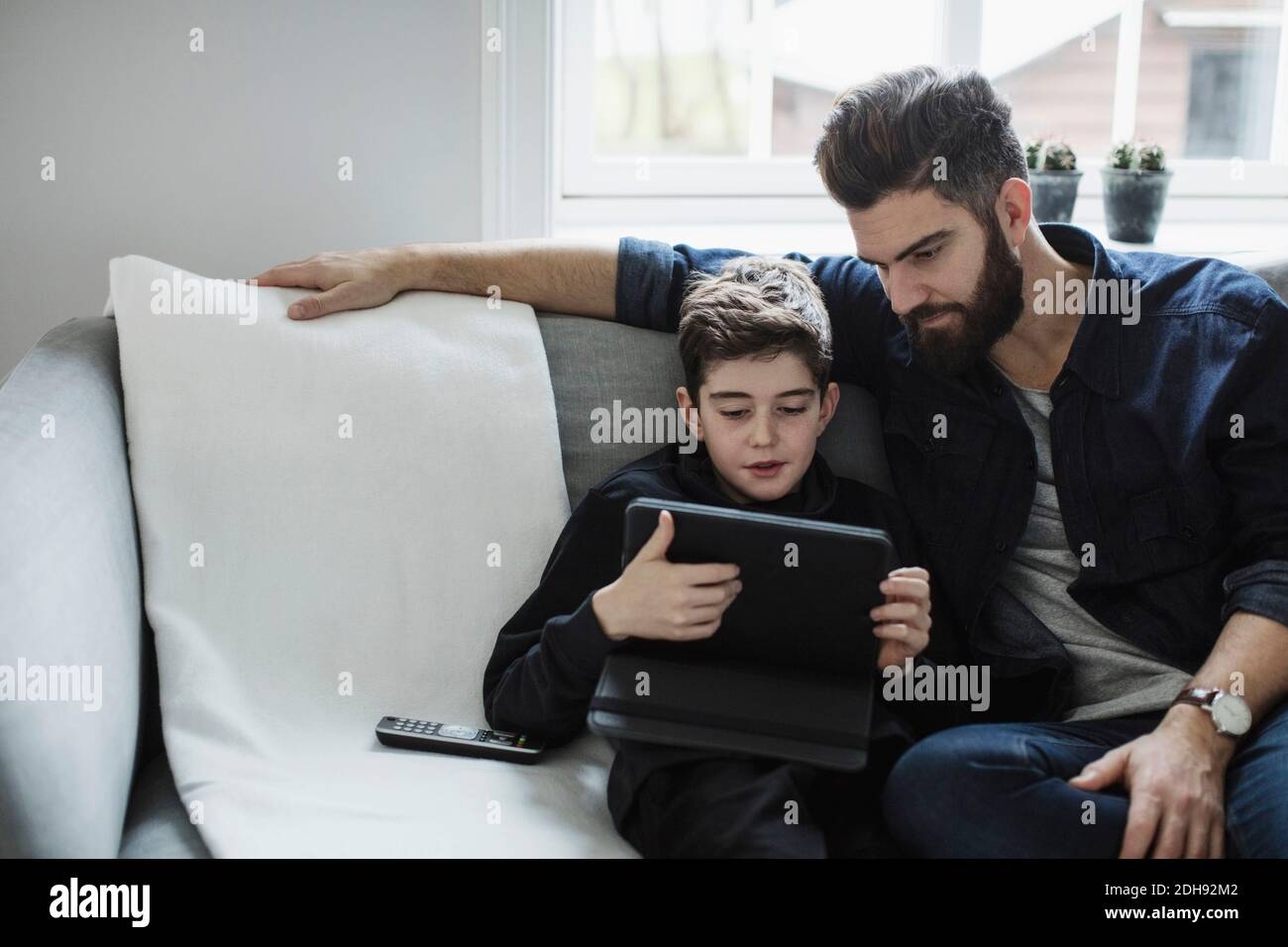 Father looking at boy using digital tablet while sitting on sofa in living room Stock Photo