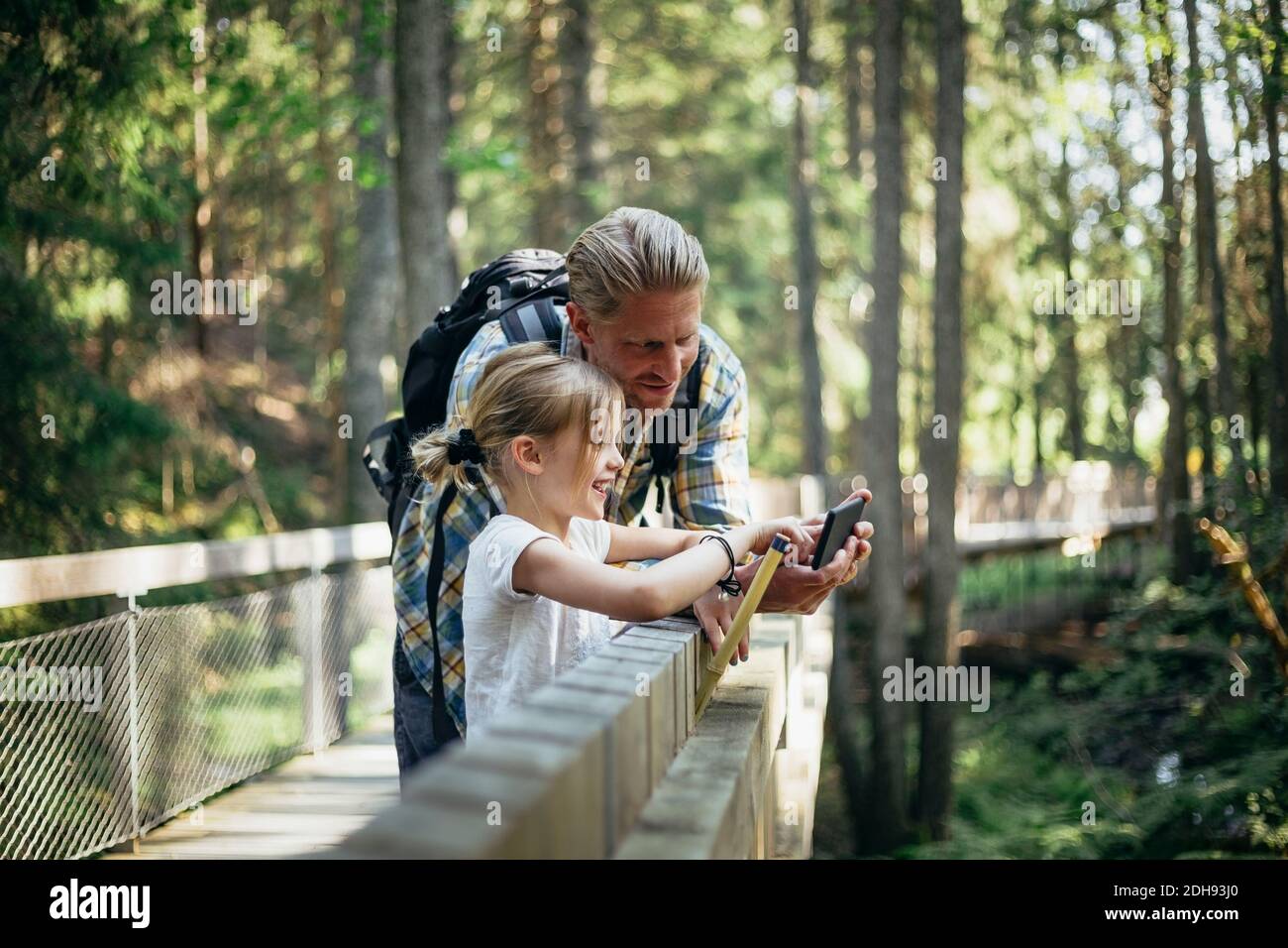 Smiling father with backpack and daughter looking at smart phone in forest Stock Photo