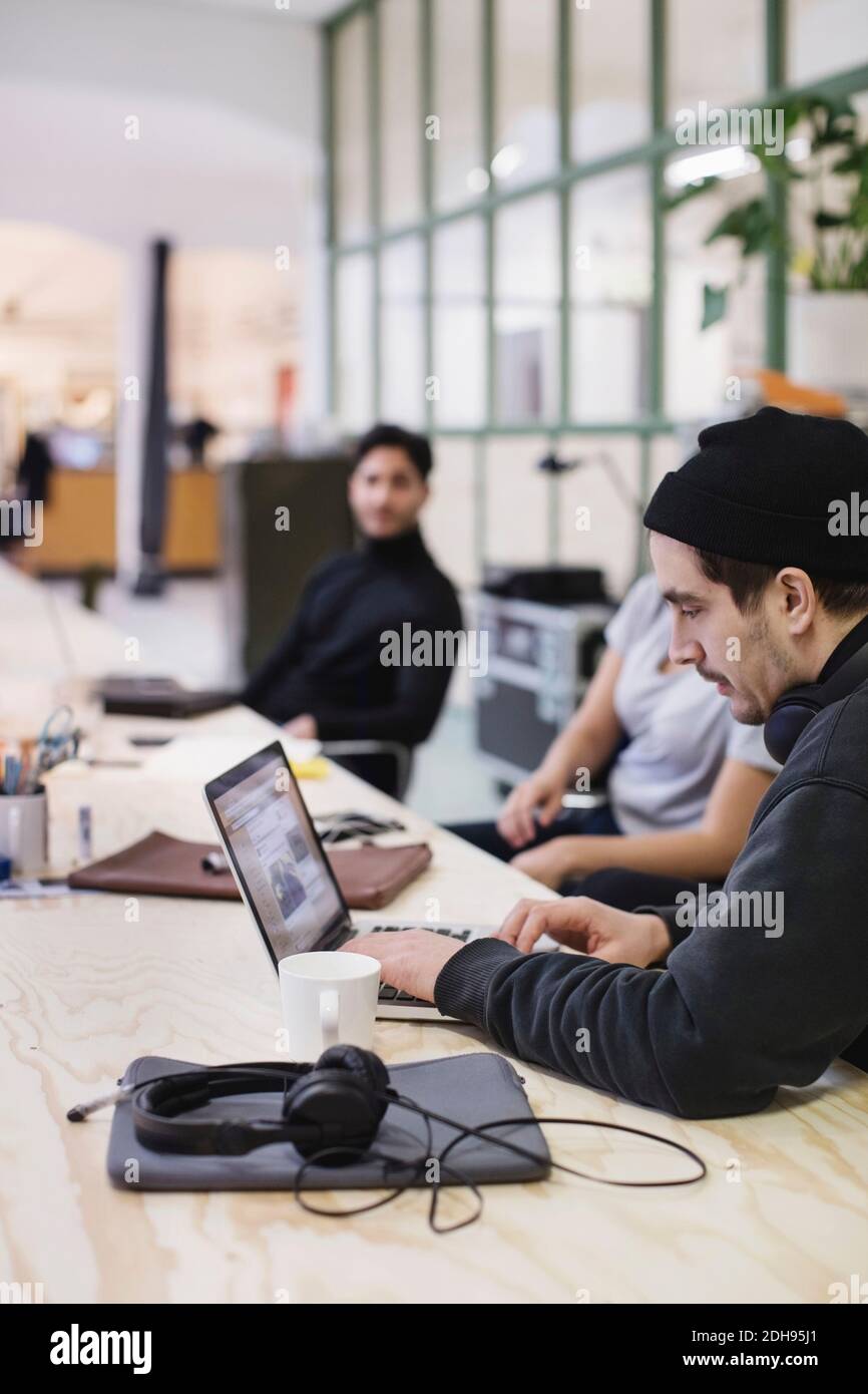 Young man using laptop at desk while colleagues sitting in background Stock Photo