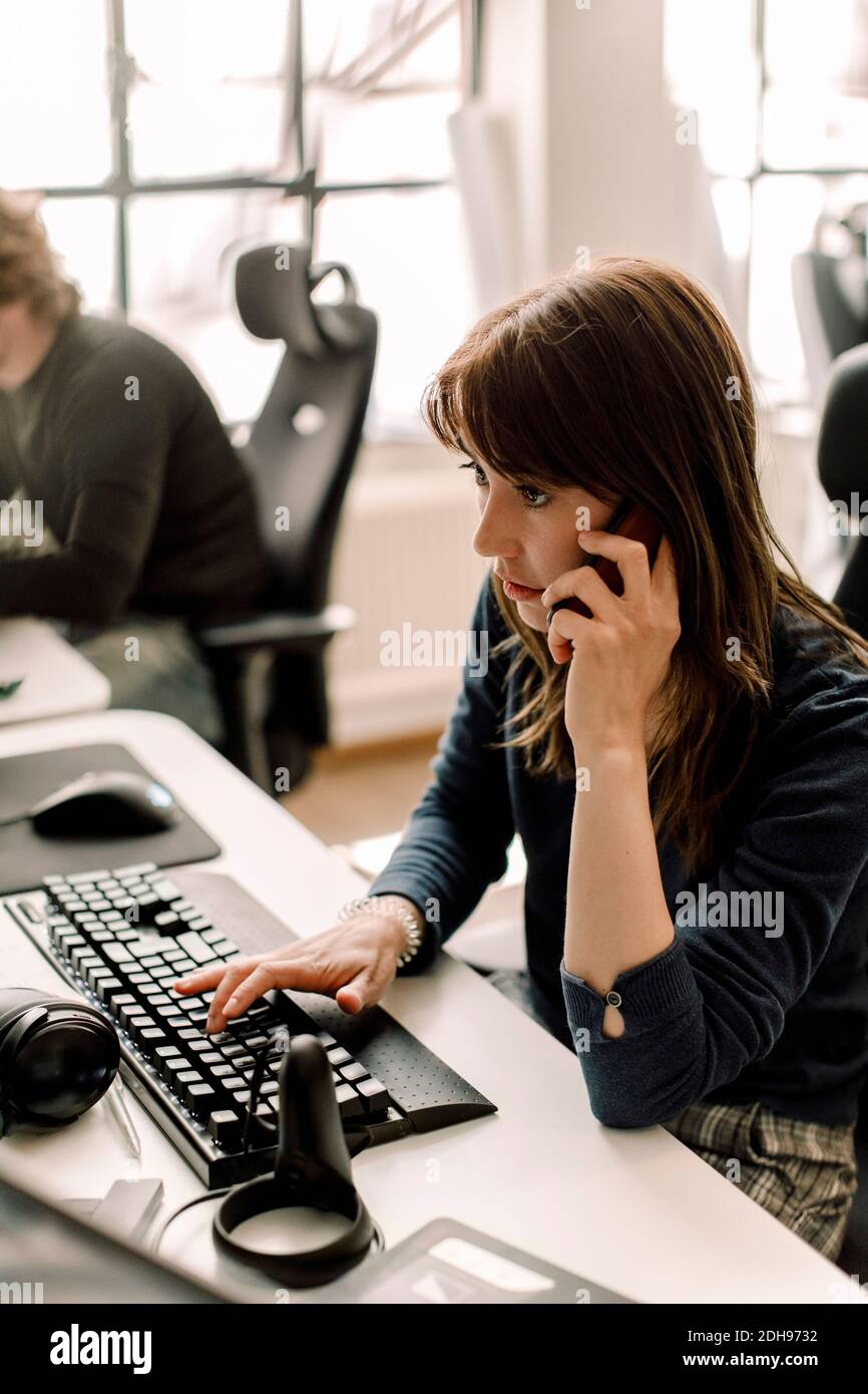 Female entrepreneur working on programming while talking on phone at workplace Stock Photo