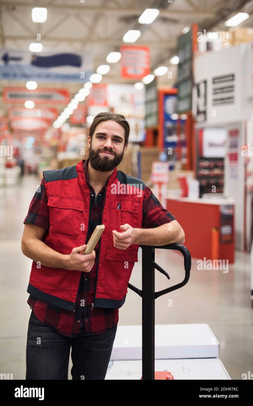 Portrait of confident salesman holding folding ruler while standing by handtruck in hardware store Stock Photo