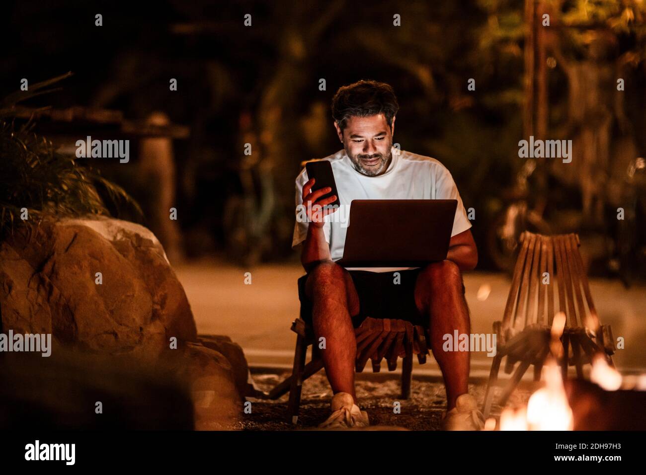 Smiling man with phone using laptop at night Stock Photo