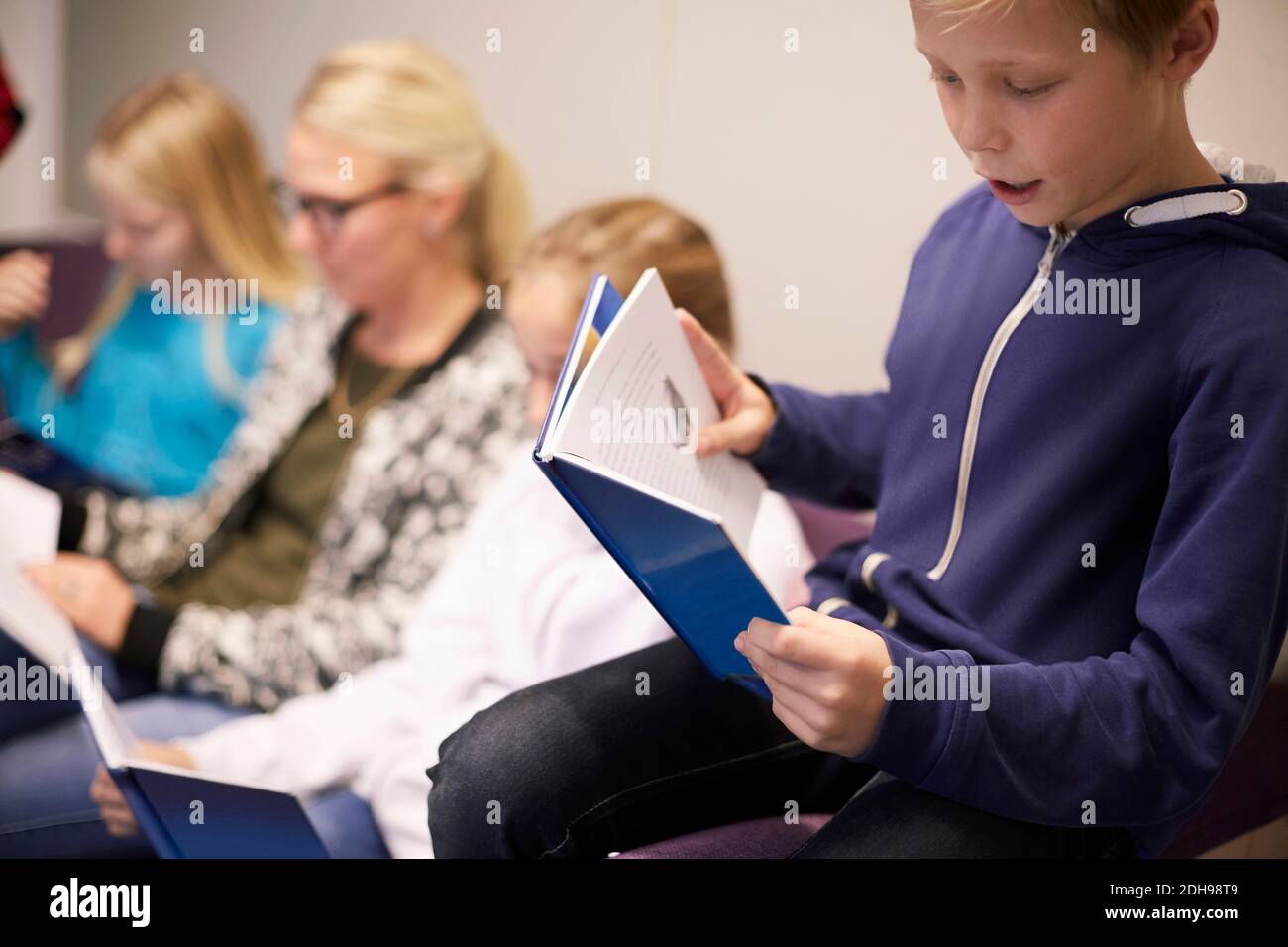 Boy reading book with friends and teacher in background at classroom Stock Photo
