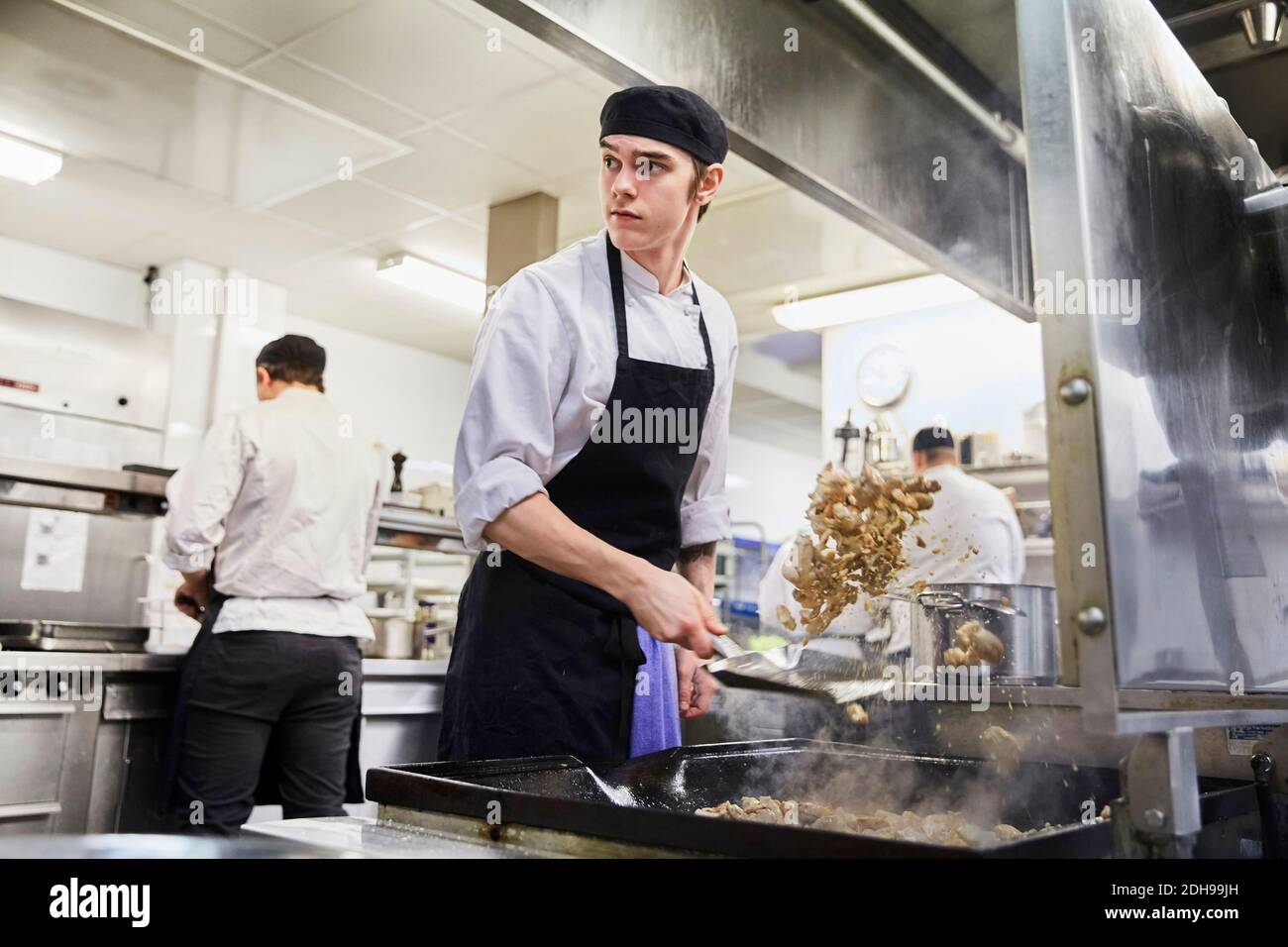 Male chef student tossing meat with colleagues in background at cooking school Stock Photo