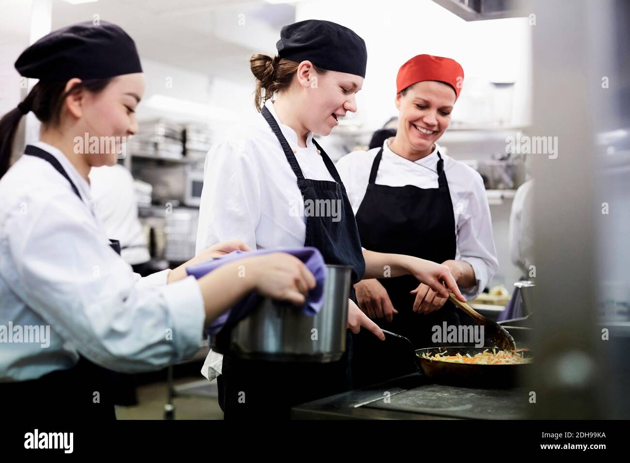 Teacher watching female chef students cooking food in commercial kitchen Stock Photo